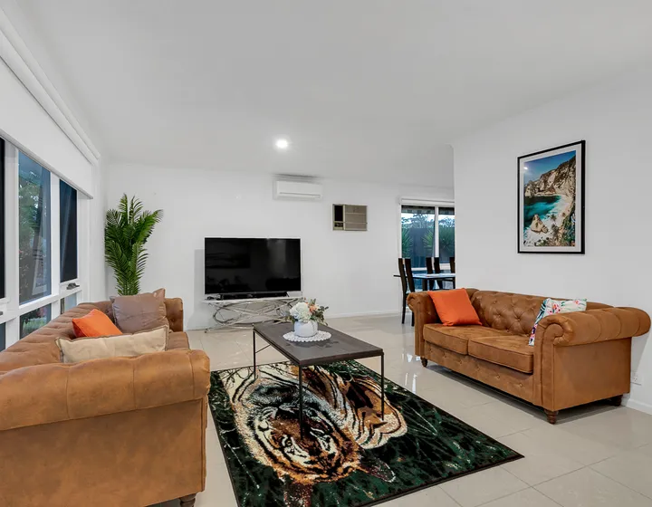 The shared living room at 3 Phelan Drive — two large leather sofas, a television and wide tiled floor, with garden windows along one wall.
